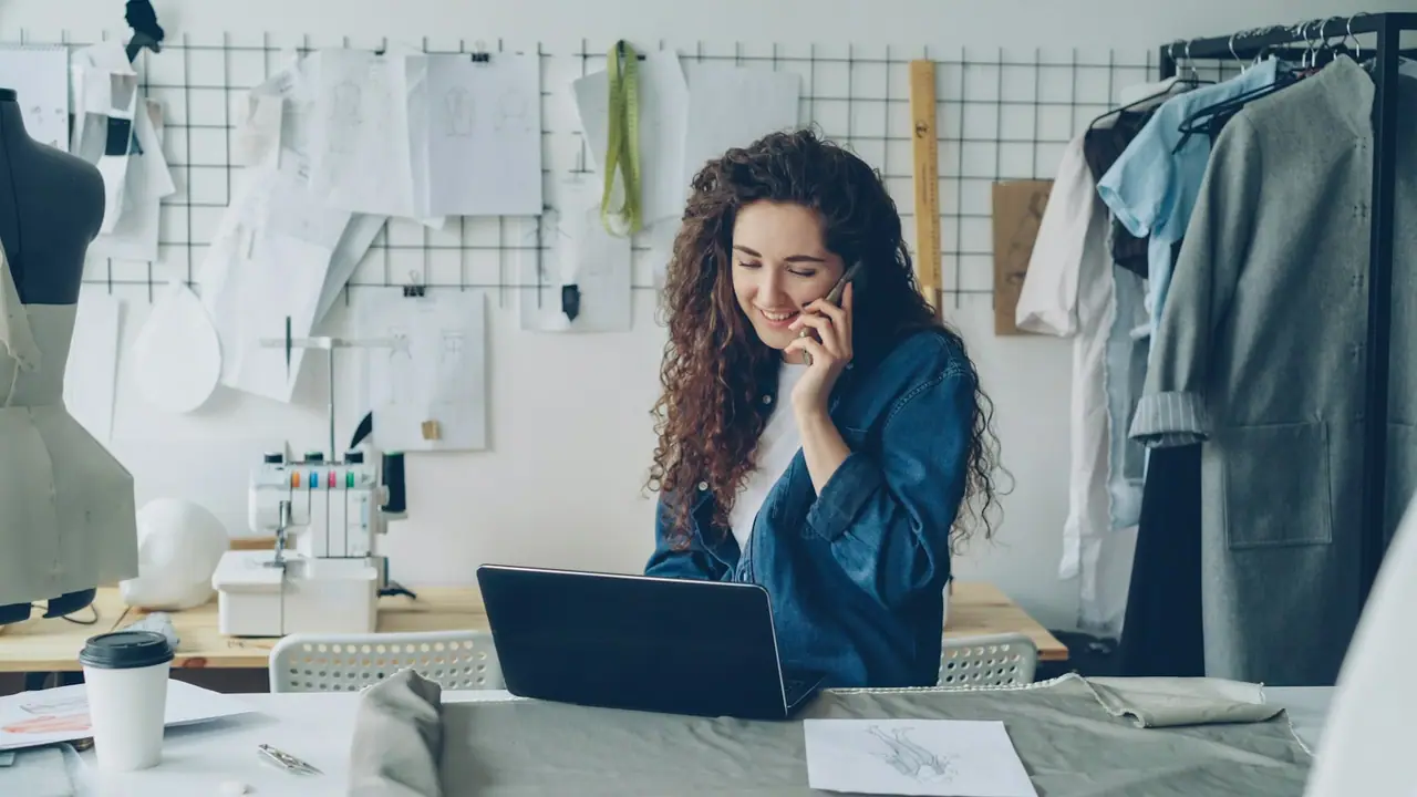 Una diseñadora habla por teléfono en su taller mientras trabaja con el portátil