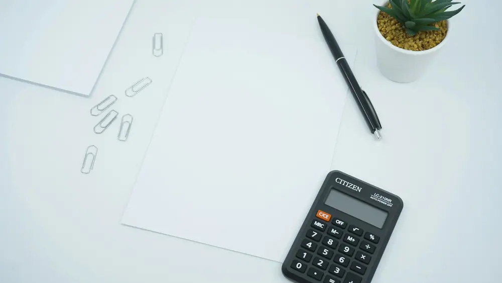Calculadora, bolígrafo y una hoja en blanco sobre una mesa blanca con una planta